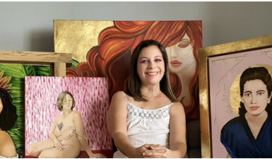 Photo of Susan Olivera sitting in front of multiple paintings, she is wearing a white t shirt, has short bob length hair, and is smiling