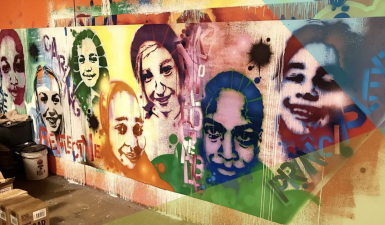 A image of a warehouse space, with a long plywood canvas with spray painted images of multi-colored faces of happy people