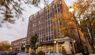 A photo of the uptown artspace lofts in Indiana. It is a 6-floor historic brick building with large windows on the outside, and large store front windows at the bottom of the building. The photo is taking from across the street, across from the building's parking lot. It is a calm fall day in a quiet downtown neighborhood.